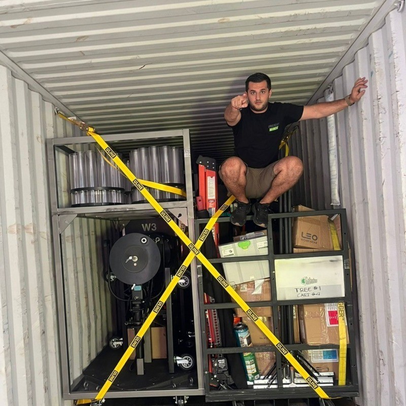 Alekos Polis crouching inside a packed shipping container, pointing forward, surrounded by securely strapped event equipment and freight boxes. Freight quote generation for commercial and entertainment logistics