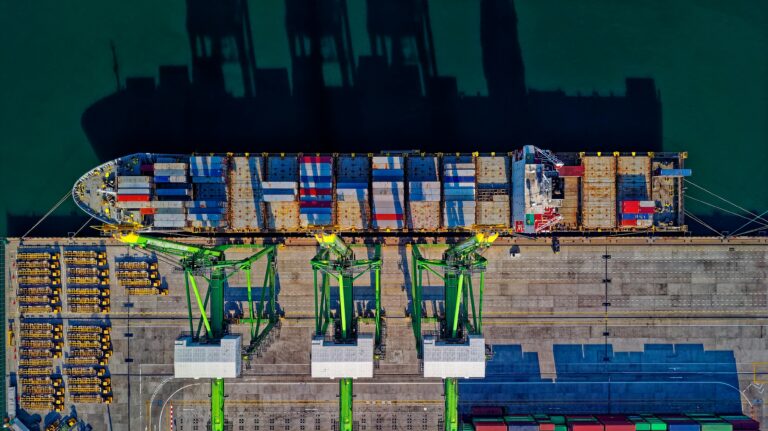 Aerial view of a cargo ship docked at a commercial port, with cranes unloading shipping containers.